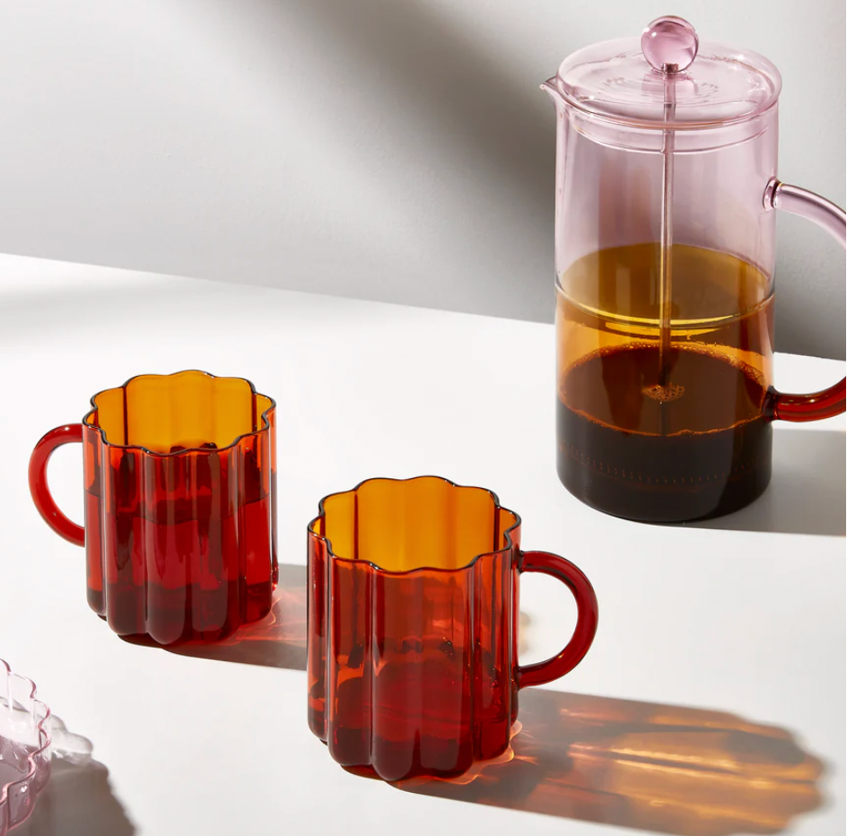 Two amber-colored mugs and a pink French press on a white surface with a cake.