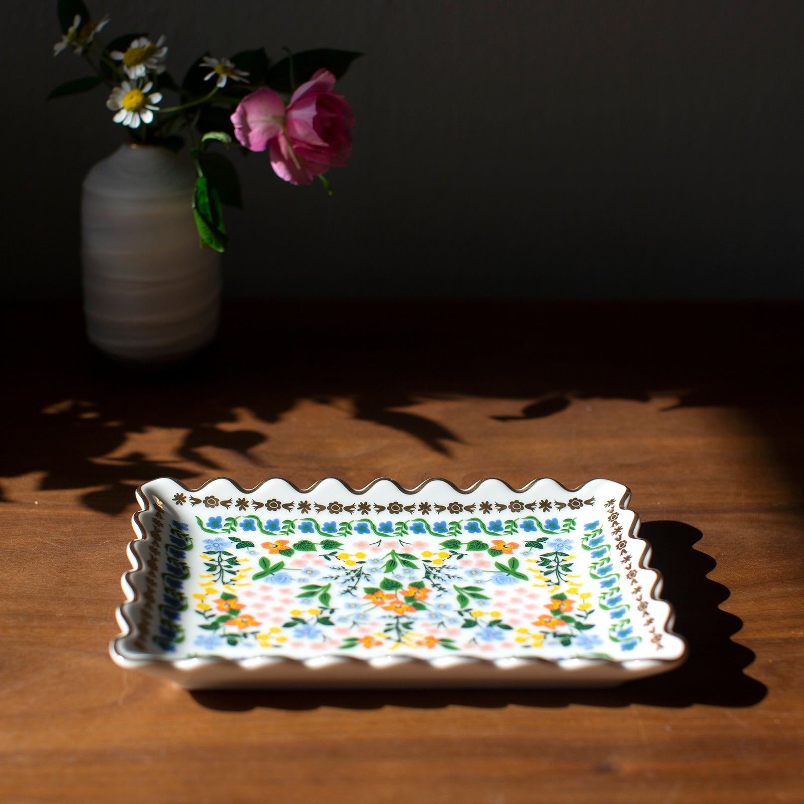 Decorative tray with floral patterns on a wooden surface with a vase of flowers in the background.